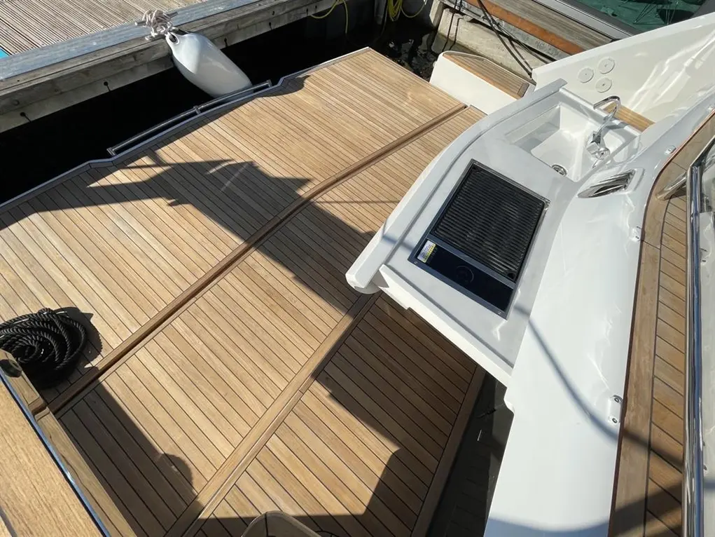 Wooden deck of a boat with a white control console, grill, and cleats in bright sunlight, showing detailed grain pattern.